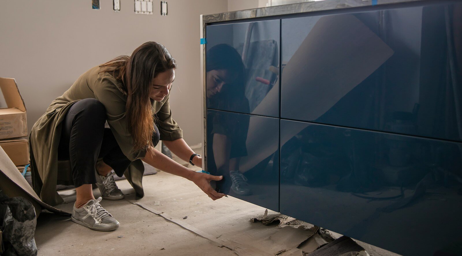 A woman with long dark hair crouching down next to a new kitchen unit being installed