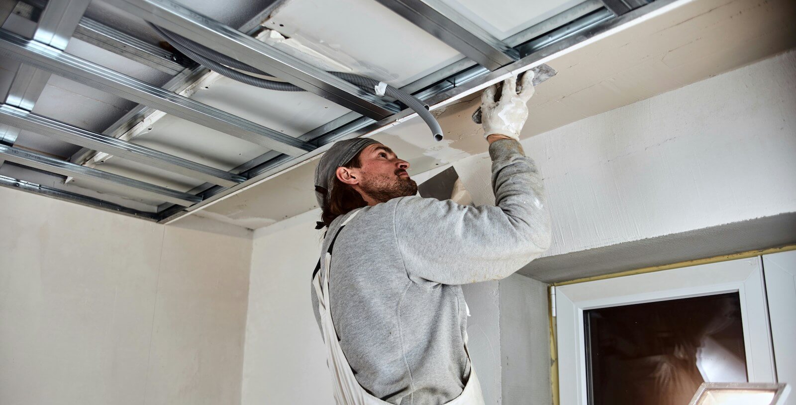 A man in white overalls up a ladder fixing steel beams to a white ceiling