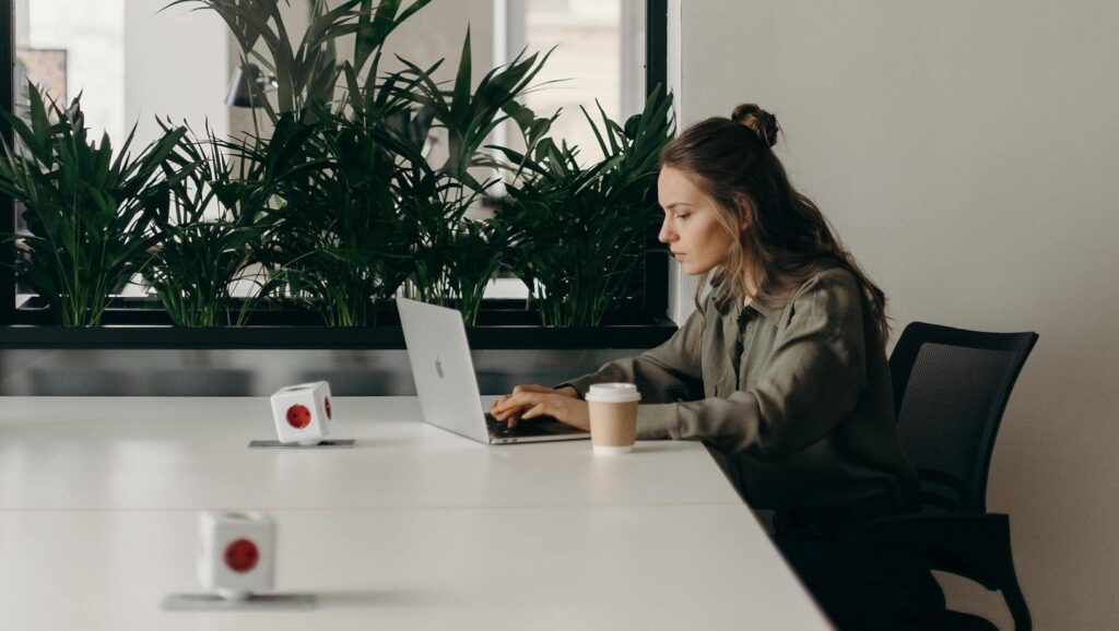 Young woman with blonde hair using a laptop on a white desk with a brown takeaway coffee cup next to indoor plants