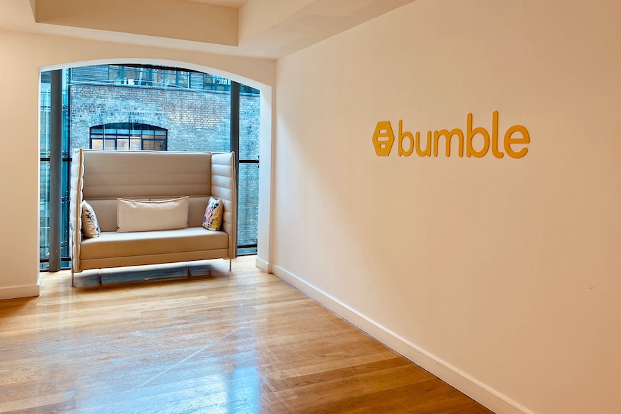 White wall with Bumble logo on it with a floor to ceiling window and grey soft seat