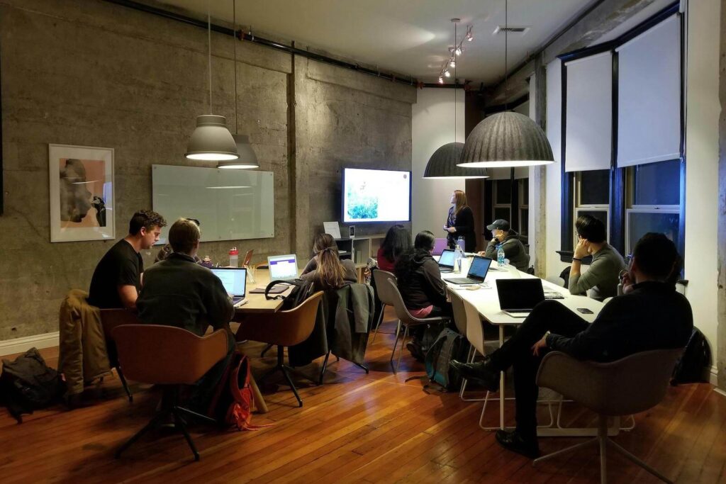 Group of people looking at a screen in an office meeting room with concrete walls, glass doors and dark wood floor