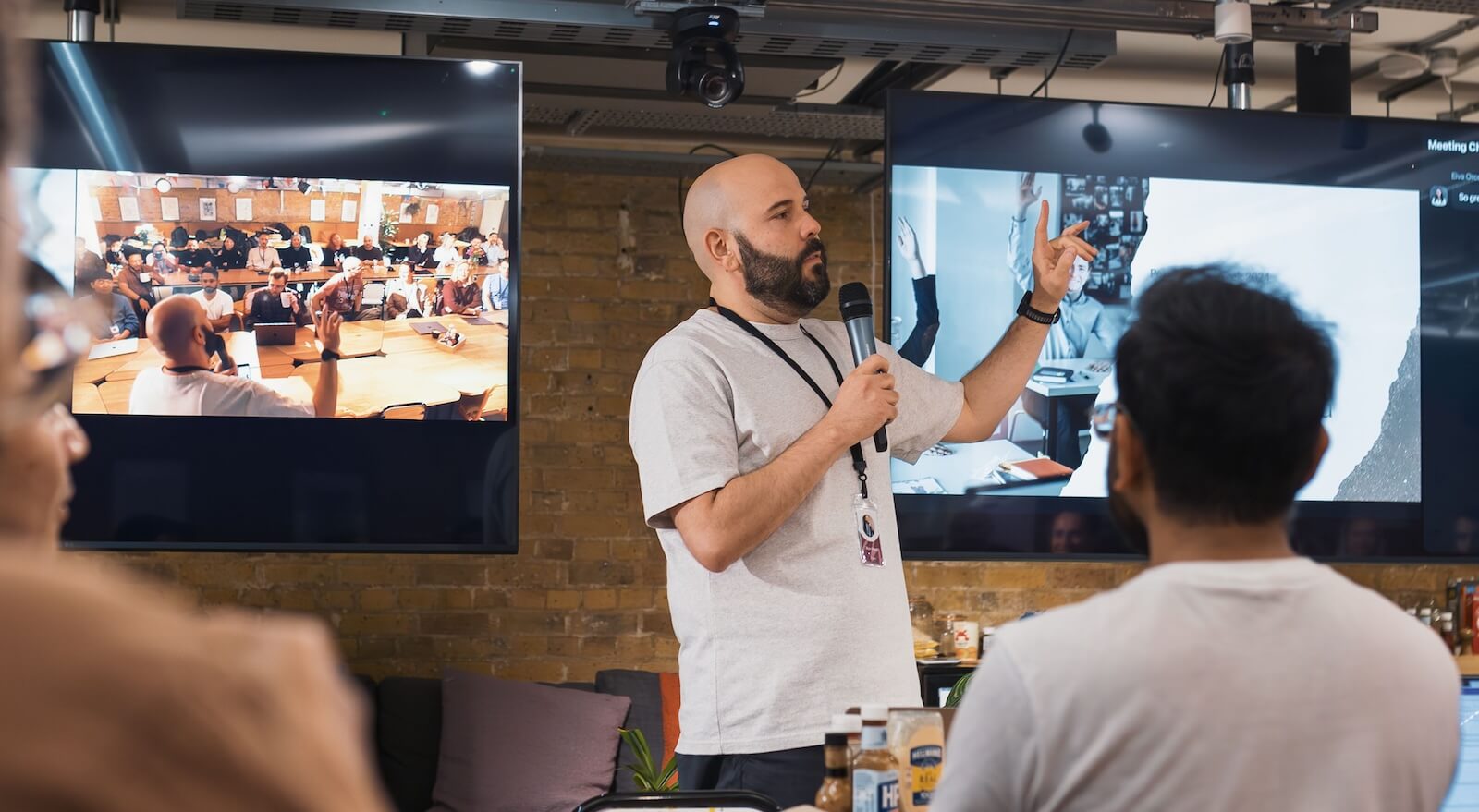 A man with a microphone talking to a room of people with two screens either side of him with people dialling in via video call